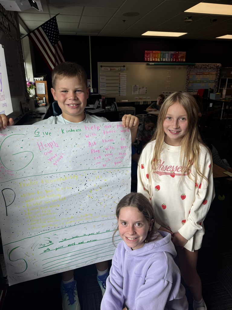 Three elementary students in a classroom smile while holding up a handwritten poster labeled "GPS" with rules for kindness, accountability, and safety.