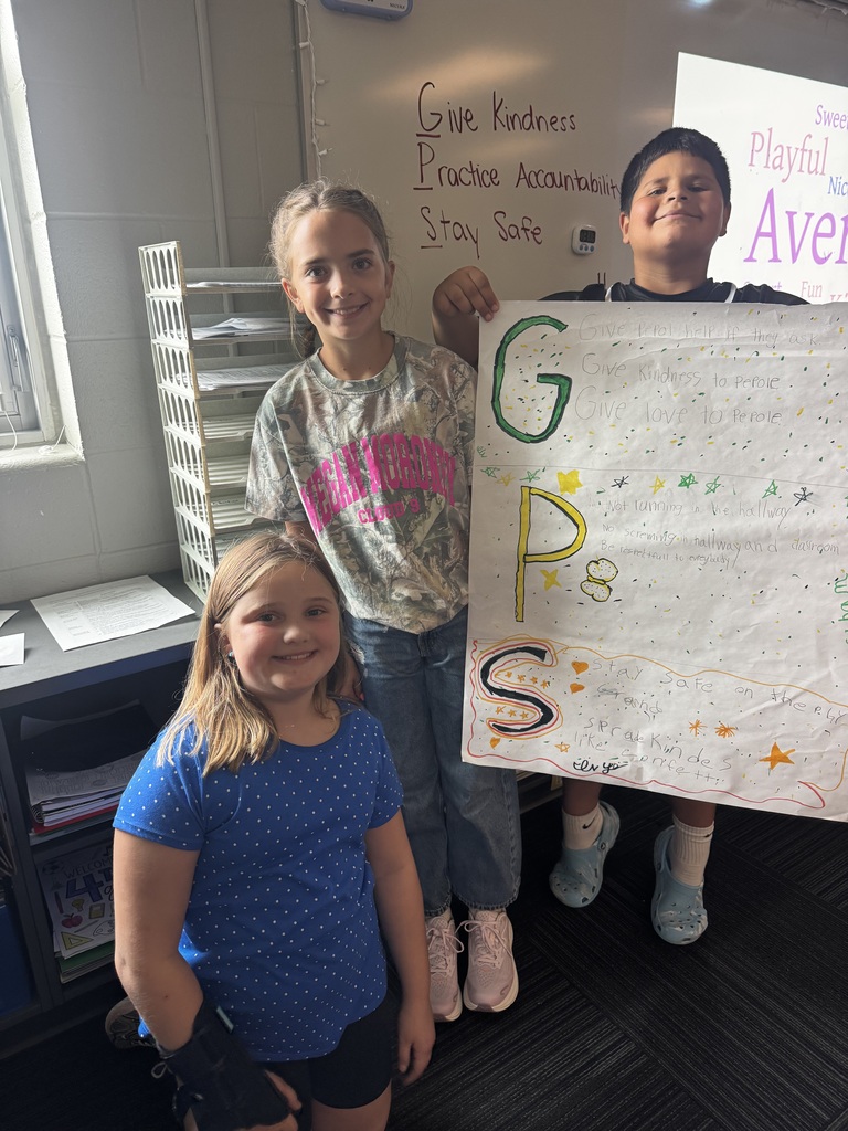 Three young students pose together in a classroom, with one boy holding up a large poster outlining "GPS: Give Kindness, Practice Accountability, Stay Safe."