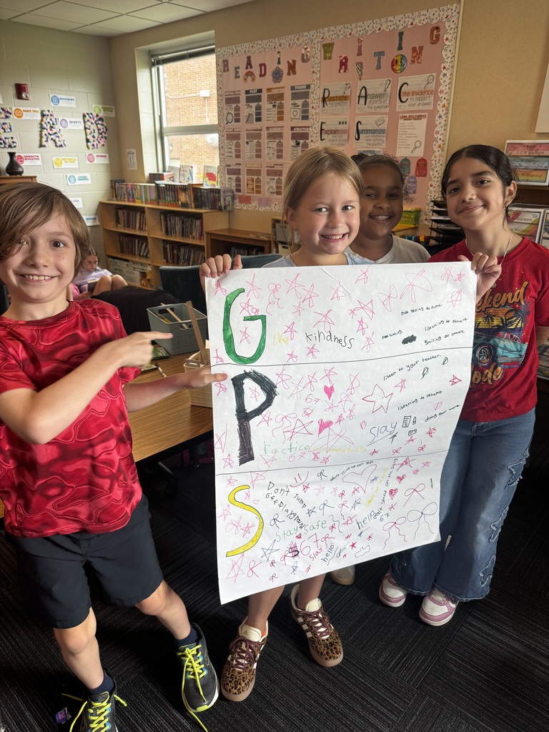 Four young students stand in a classroom around a large poster covered in star drawings that reads "GPS: Give Kindness, Practice Accountability, Stay Safe."