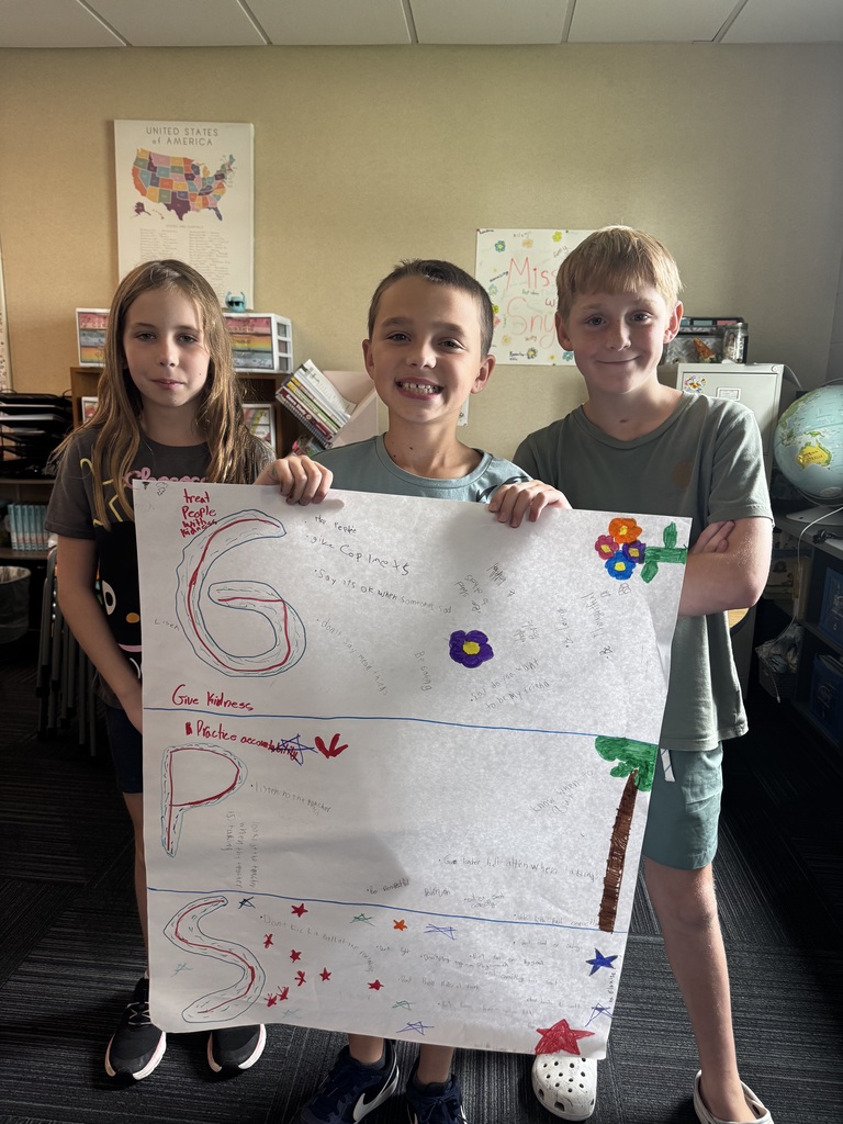 Three elementary school students stand together in a classroom, holding a large poster decorated with drawings and notes explaining "GPS" school guidelines.