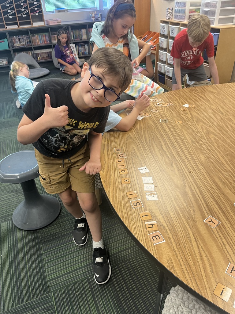 A boy wearing glasses and a Jurassic World shirt gives a thumbs-up next to a wooden table with letter tiles.