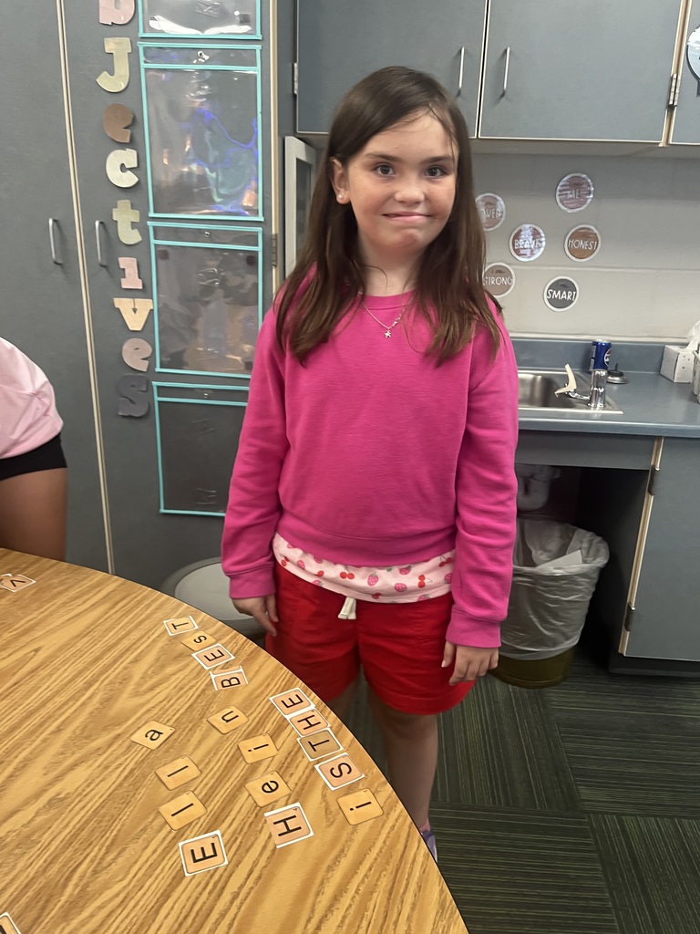 A young girl in a bright pink sweater and red shorts stands near a table with letter tiles spelling out a message.