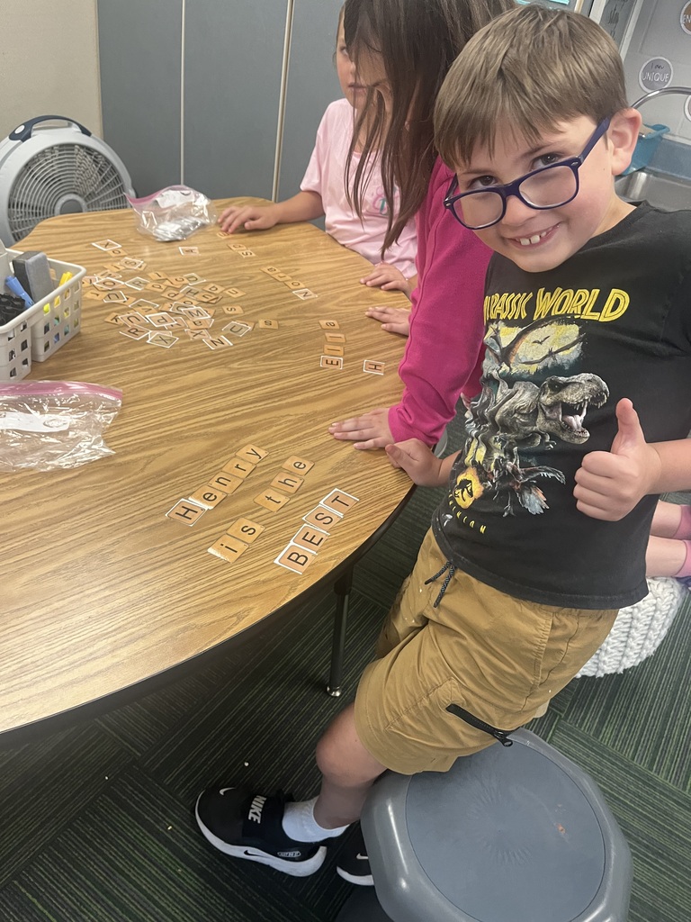 A young boy in a Jurassic World t-shirt gives a thumbs-up while standing next to a table where letter tiles spell "Henry is the BEST".