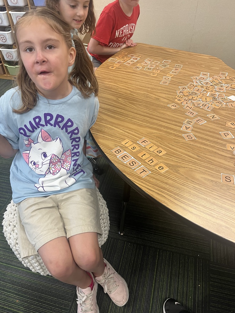 A young girl in a light blue cat-themed t-shirt sits on a pouf next to a table displaying letter tiles.
