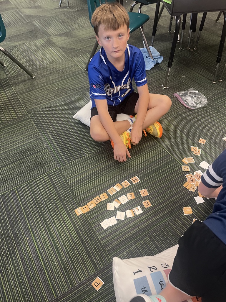 A boy wearing a blue sports jersey sits cross-legged on a pillow on the floor near letter tiles arranged on the carpet.