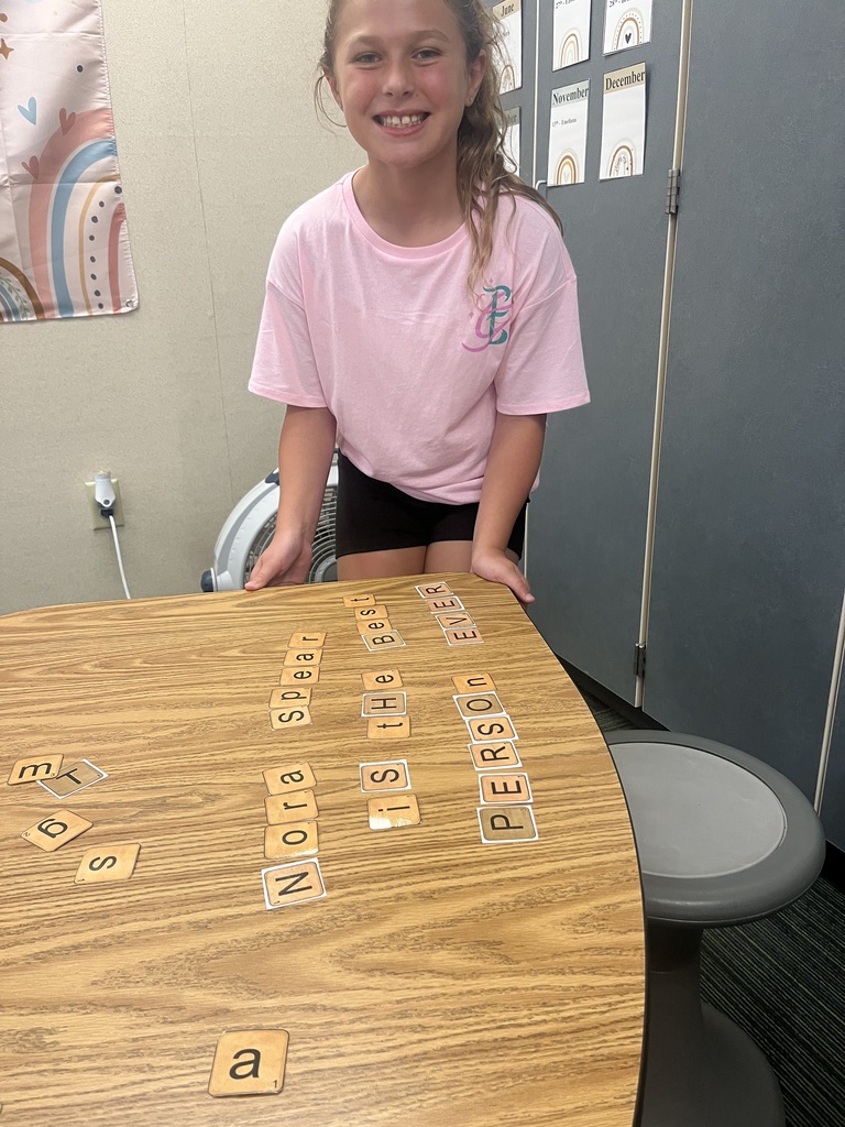 A smiling girl in a light pink shirt stands at a table holding letter tiles that spell out "Nora Spear is the BEST PERSON EVER".