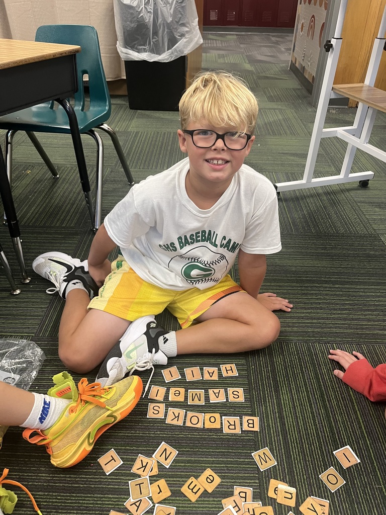 A young boy wearing glasses and a baseball shirt sits on a classroom floor smiling near letter tiles arranged into words.