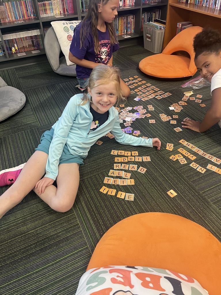 A blonde girl smiles at the camera while sitting on the floor surrounded by scattered and arranged letter tiles.