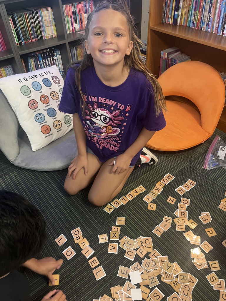 A smiling girl in a purple shirt kneels on a classroom carpet surrounded by letter tiles that spell out various words and names.