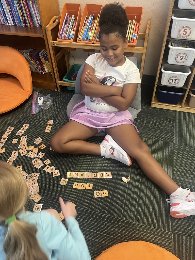 A young girl sits crossed-legged on the floor with her arms crossed, smiling down at spelling tiles laying in front of her.