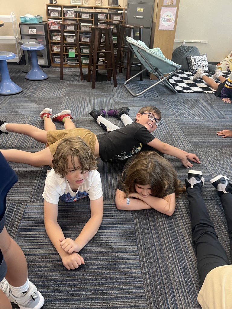 Three students lay on their stomachs on the carpeted floor, resting their arms and looking toward the camera.