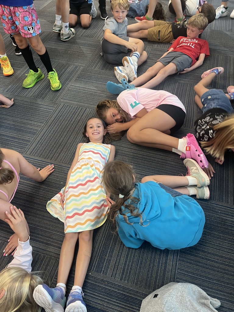A young girl in a colorful striped dress lies on the carpet and smiles, surrounded by classmates sitting and lying down around her.