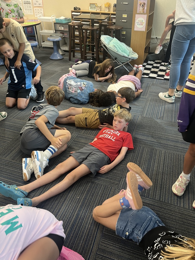 Several elementary students lie down on the classroom carpet in different directions during an indoor activity.