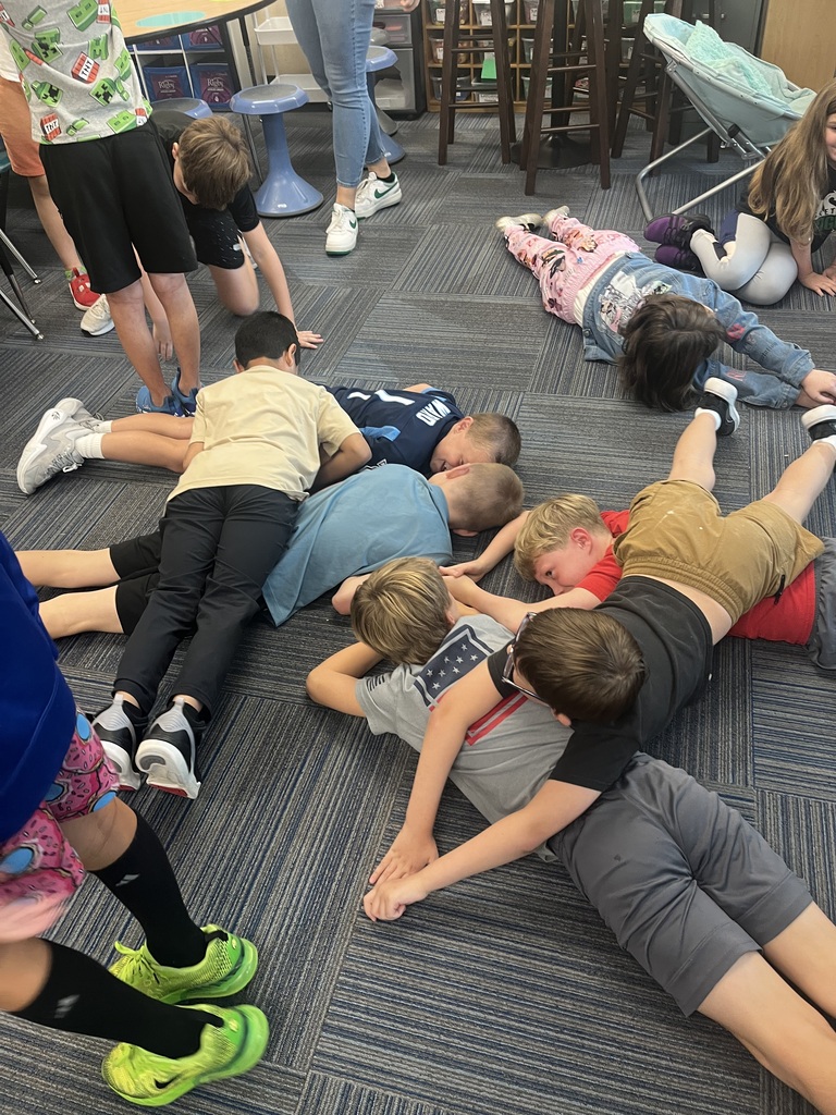 A group of young students lay face down on the classroom floor, positioning themselves closely together as part of an exercise.