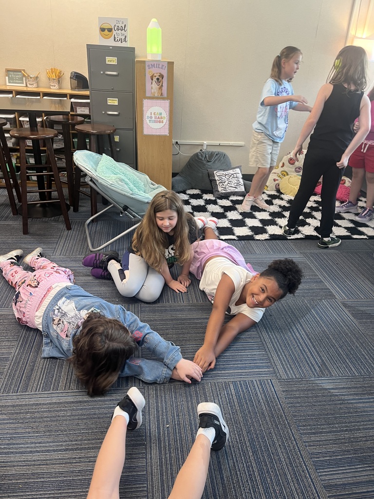 Three girls lay on the carpet reaching toward each other and smiling while classmates play nearby in the background.