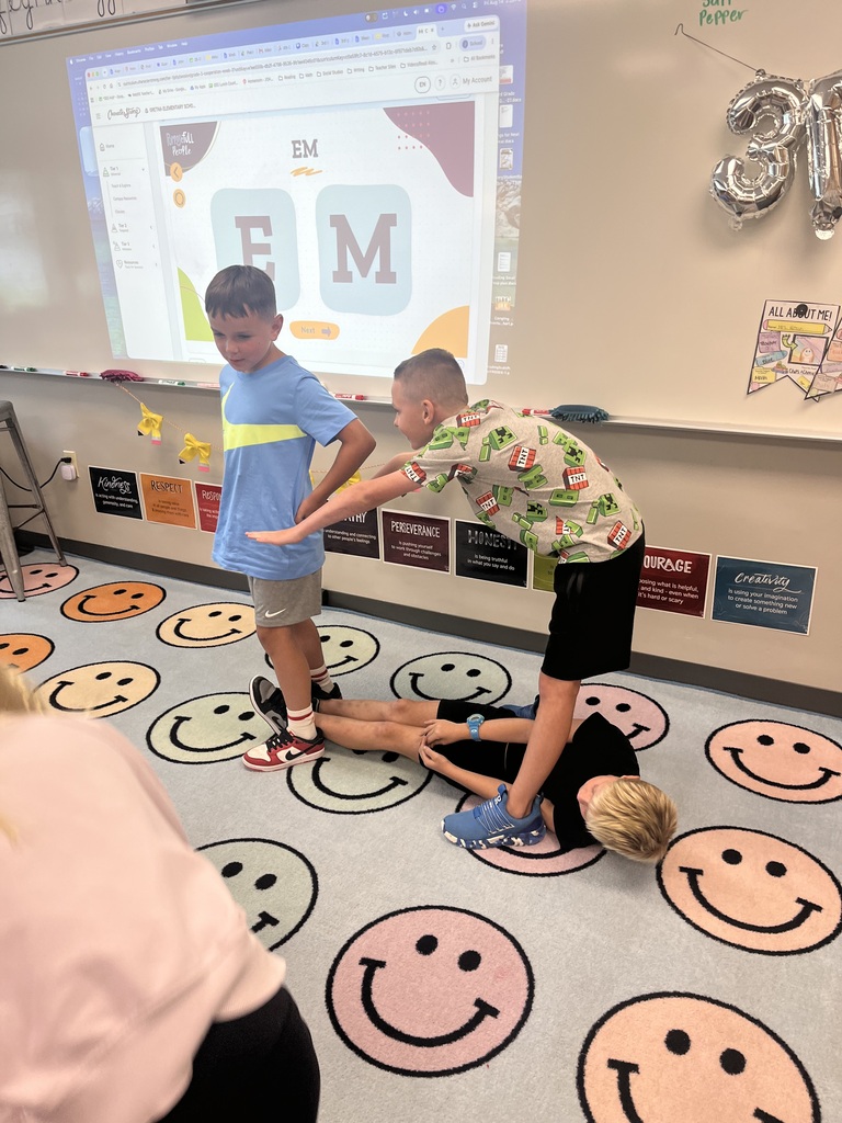 Three young boys pose together on a classroom carpet decorated with smiley faces while participating in a group activity.