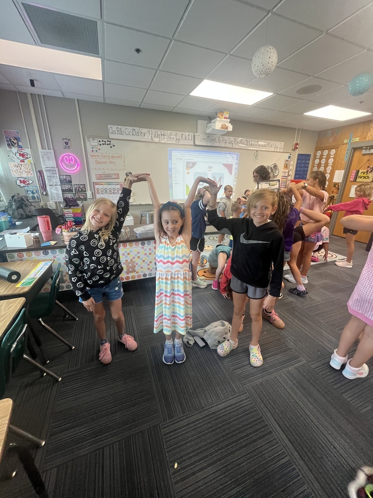 Three young girls hold hands above their heads to form shapes in a classroom, with other students practicing similar poses in the background.