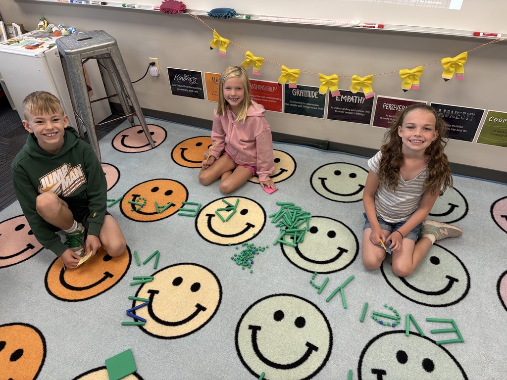 Three young students smiling on a classroom rug with smiley faces, using green base-ten blocks to spell out names.