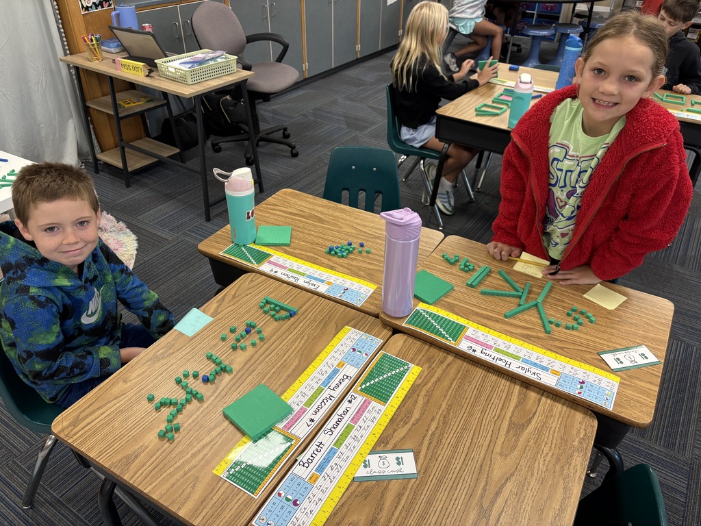 Two elementary students sitting at adjoining wooden desks in a classroom, posing with green math manipulatives spelling their names.