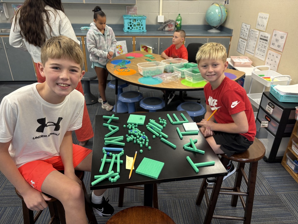 Two smiling boys sitting at a small black table covered in green math base-ten blocks spelling out "Xander and Tate".