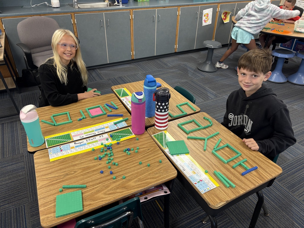 A girl and a boy at classroom desks smiling behind their names spelled out using green base-ten blocks.