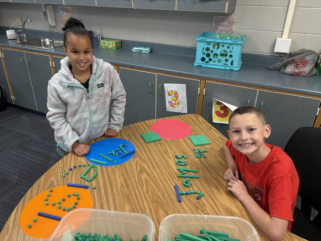 A young girl and boy smiling at a wooden table with colorful placemats and names spelled with green counting cubes.