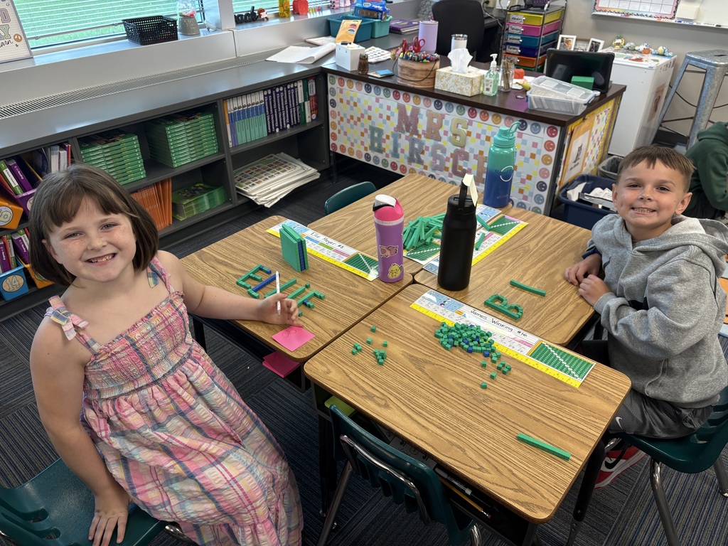 A young girl in a plaid dress and a boy in a gray hoodie sitting at desks with base-ten block activities.