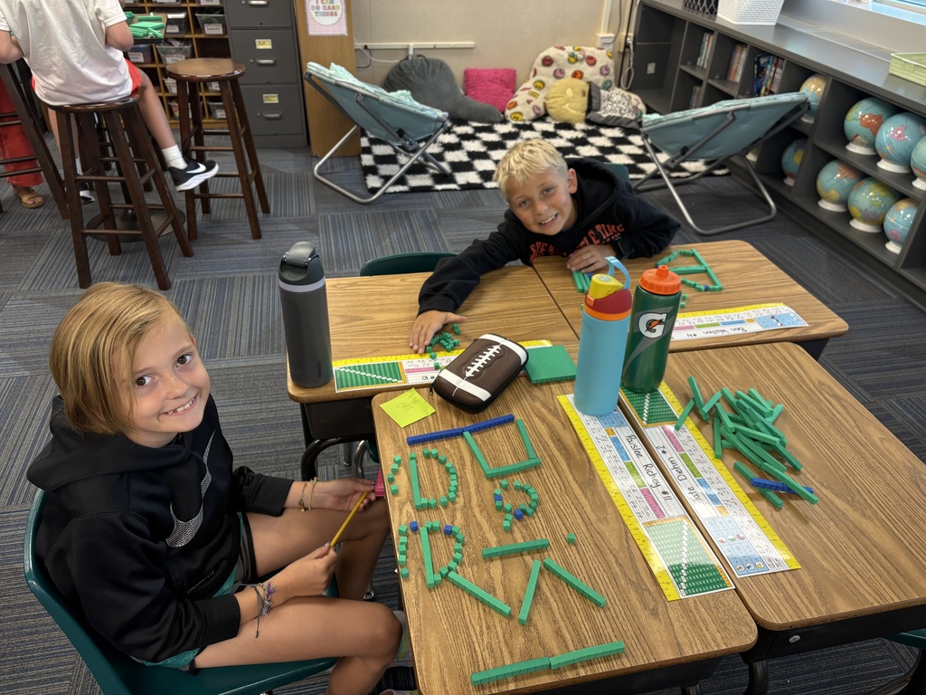 Two boys at classroom desks smiling while creating words out of green math counting blocks.