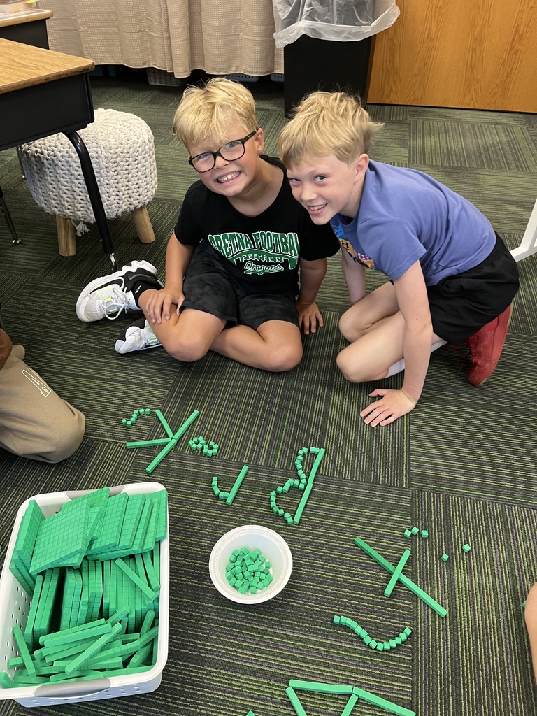 Two young boys kneel together on the carpet and smile next to green base ten blocks arranged on the floor.