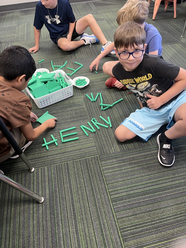 A boy wearing glasses and a Jurassic World shirt gives a thumbs-up while sitting near green base ten blocks spelling the name "Henry" on the carpet.