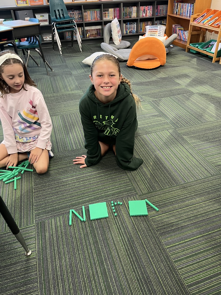A young girl in a green hoodie smiles while kneeling on a classroom carpet behind the name "Nora" spelled out with green base ten block manipulatives.
