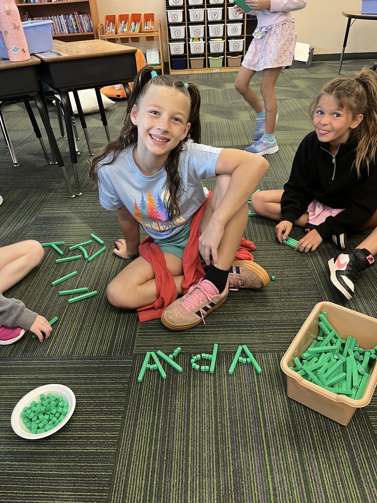 A girl in a light blue shirt smiles while sitting on the floor next to the name "Ayla" spelled out with green base ten blocks.