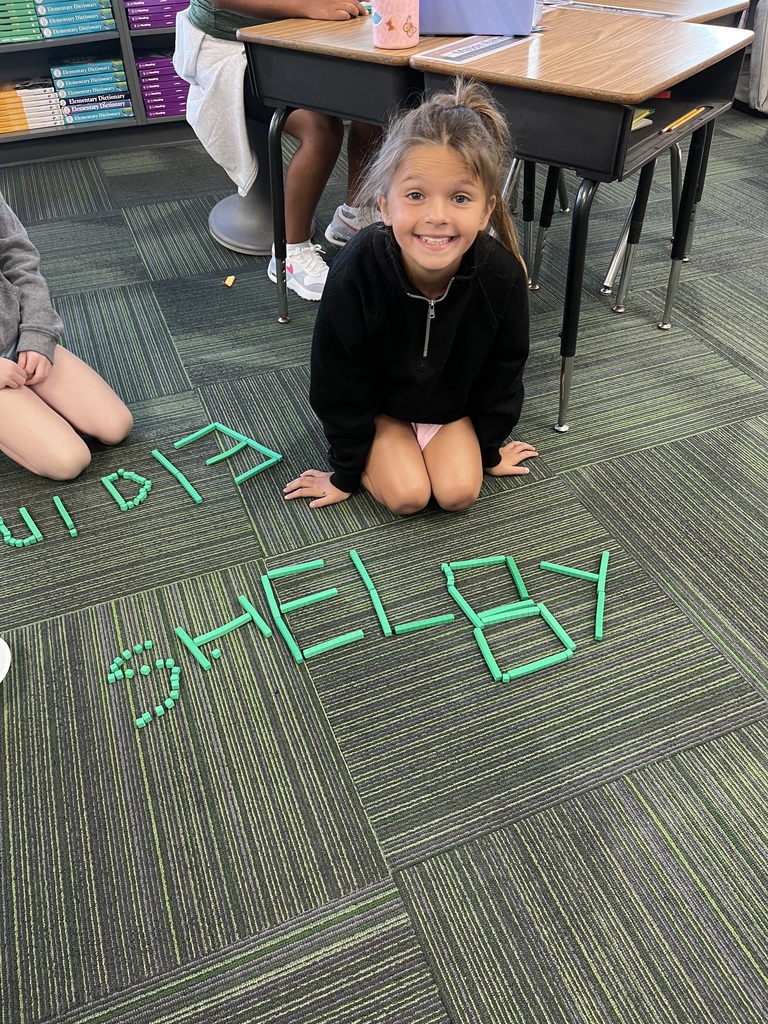 A young girl smiles brightly while kneeling on the carpet behind her name, "Shelby," created using green base ten blocks.