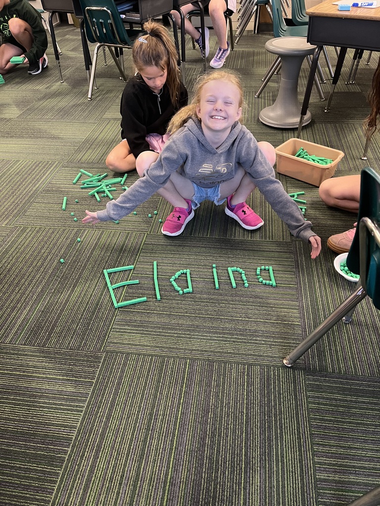 A smiling young girl in a gray hoodie crouches low over the name "Elaina" spelled out on the floor with green base ten blocks.