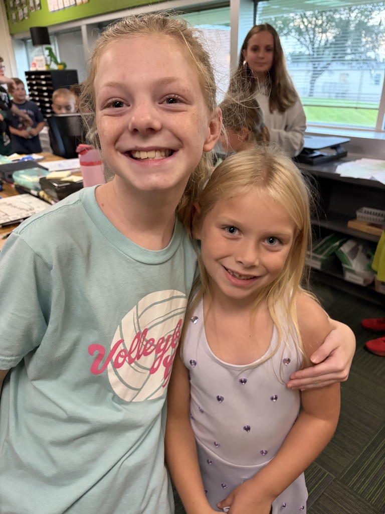 Two young girls smiling in a classroom, one in a light blue volleyball shirt with her arm around the other in a light purple dress with hearts.