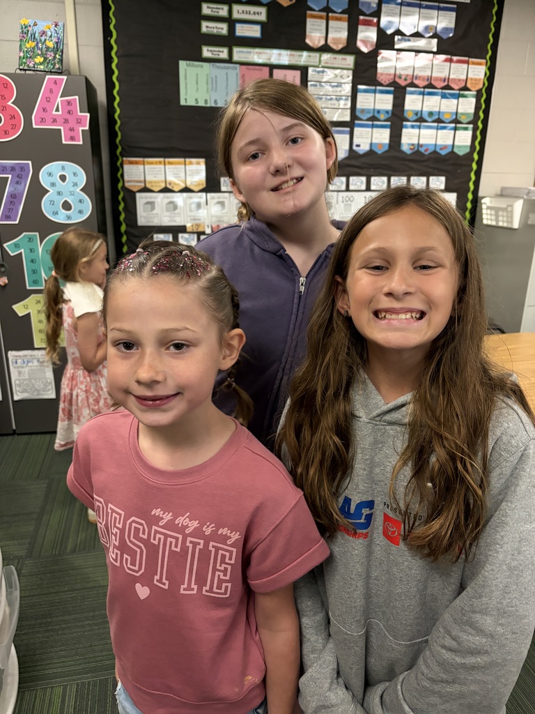 Three young girls standing together and smiling in front of a classroom math bulletin board.