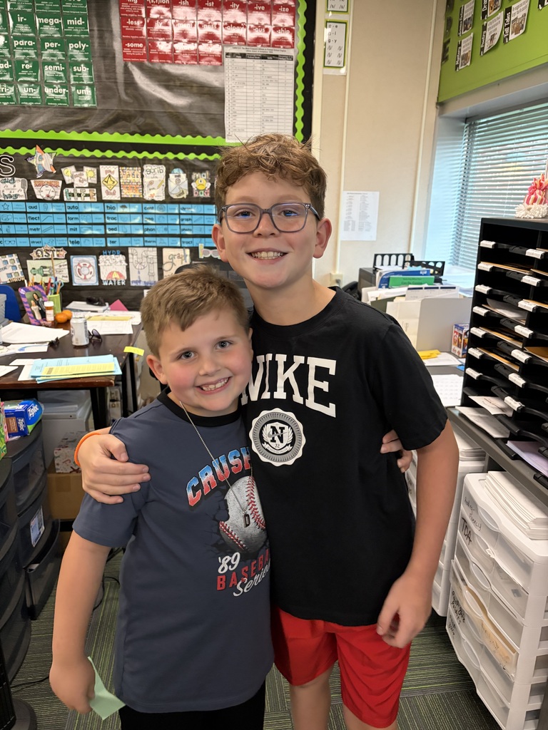Two young boys hugging and smiling, one wearing a navy baseball-themed shirt and the other in a black Nike shirt and glasses.