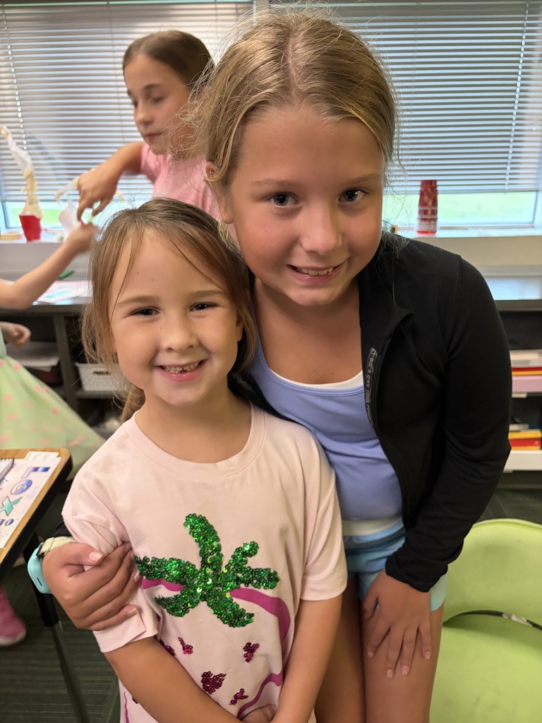 Two young girls smiling, with the taller girl in a black jacket and blue top hugging the younger girl in a pink shirt with a green sequin palm tree.
