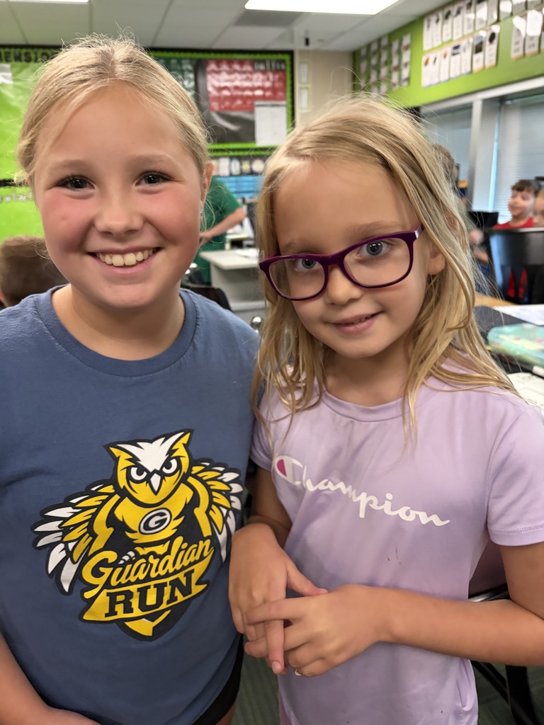Two young girls smiling side-by-side, one in a blue "Guardian Run" shirt and the other wearing glasses and a purple Champion shirt.