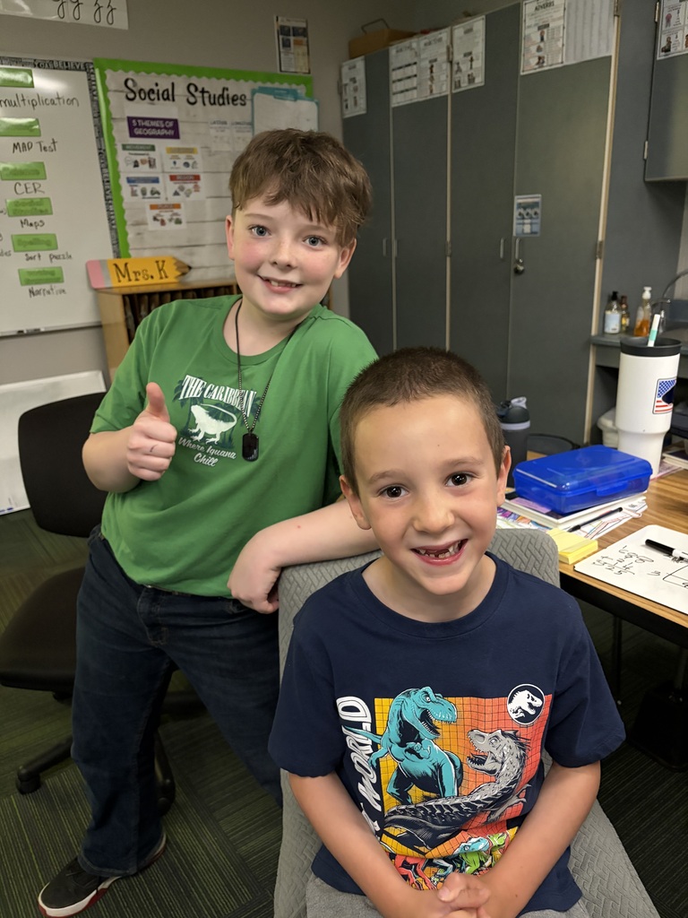 Two young boys in a classroom, one in a green shirt giving a thumbs-up while leaning over the other boy sitting in a blue dinosaur shirt.