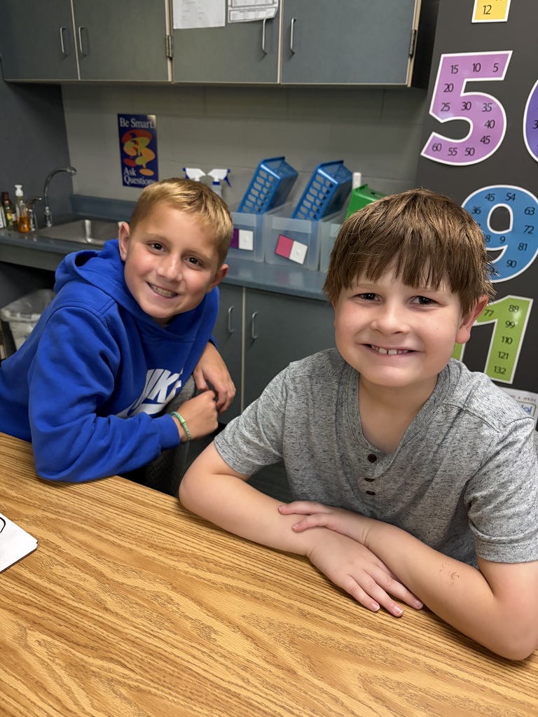 Two young boys seated at a wooden desk smiling, one in a blue Nike hoodie and the other in a grey heathered shirt.