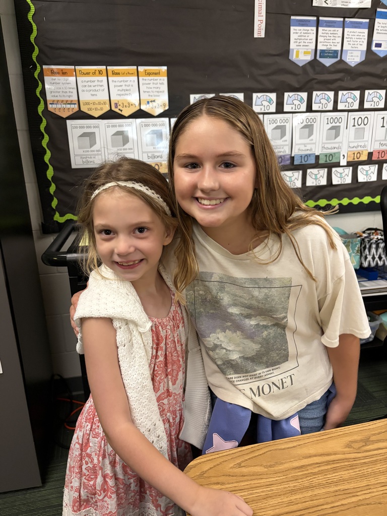 Two young girls smiling in front of a math poster, one in a pink patterned dress with a white shawl and the other in a Claude Monet t-shirt.
