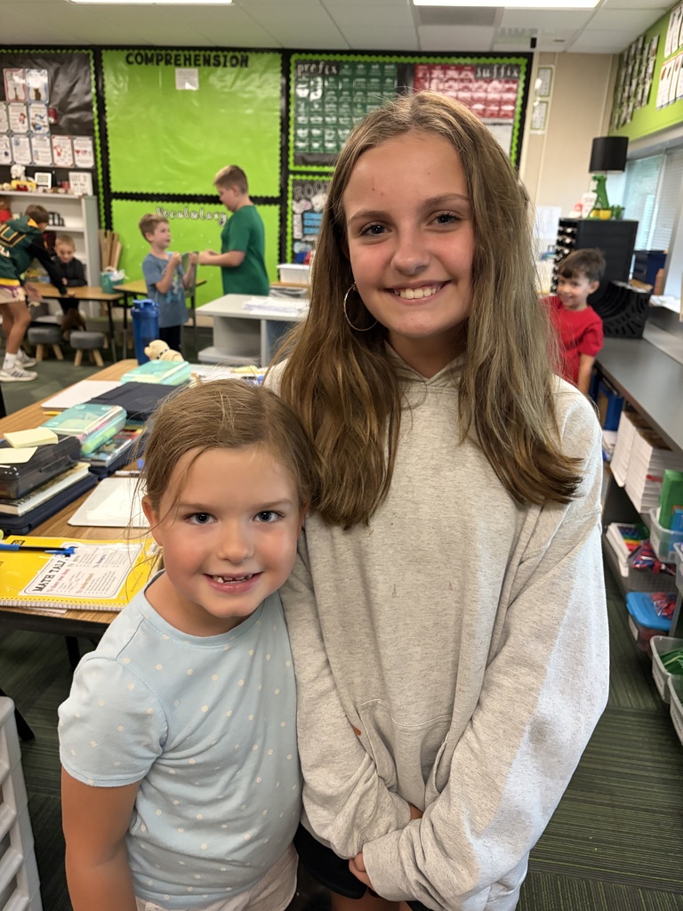 Two young girls smiling in a classroom, the taller in a grey oversized hoodie and the younger in a light blue polka-dot shirt.