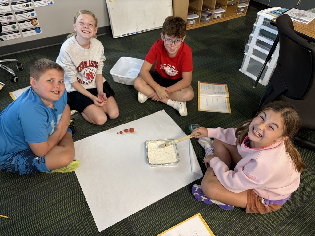 Four students sitting in a circle on the floor working on a science experiment involving a pan of flour and small clay spheres set up on paper.