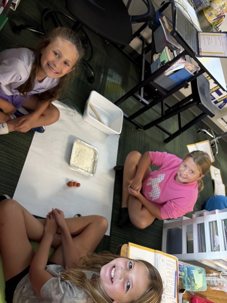 Three girls sitting in a circle on the floor smiling while gathered around a classroom science setup with flour and clay spheres.