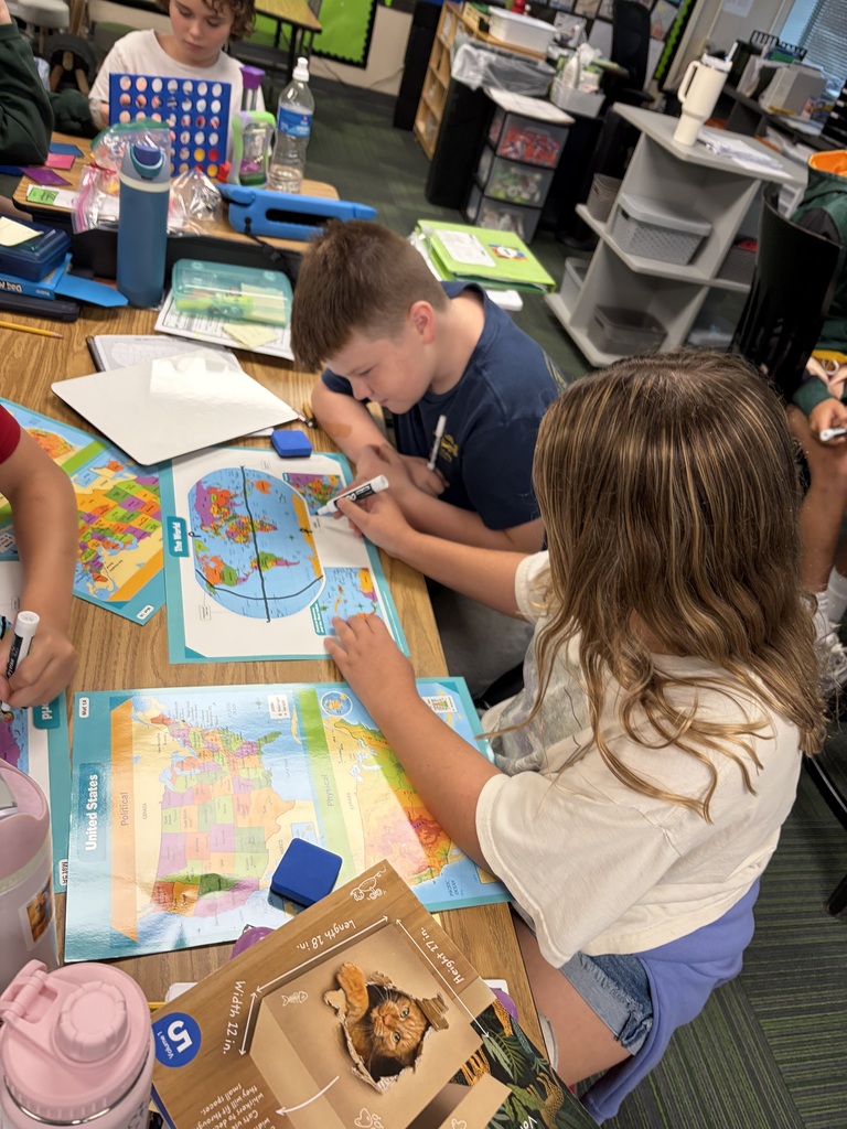 A boy in a blue shirt and a girl with long wavy hair sitting side by side at a table, marking lines on world and US map laminates with dry-erase markers.