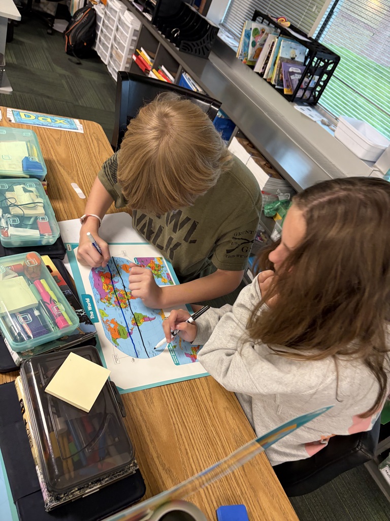 Two students leaning over a desk while tracing longitude and latitude lines onto a world map laminate.