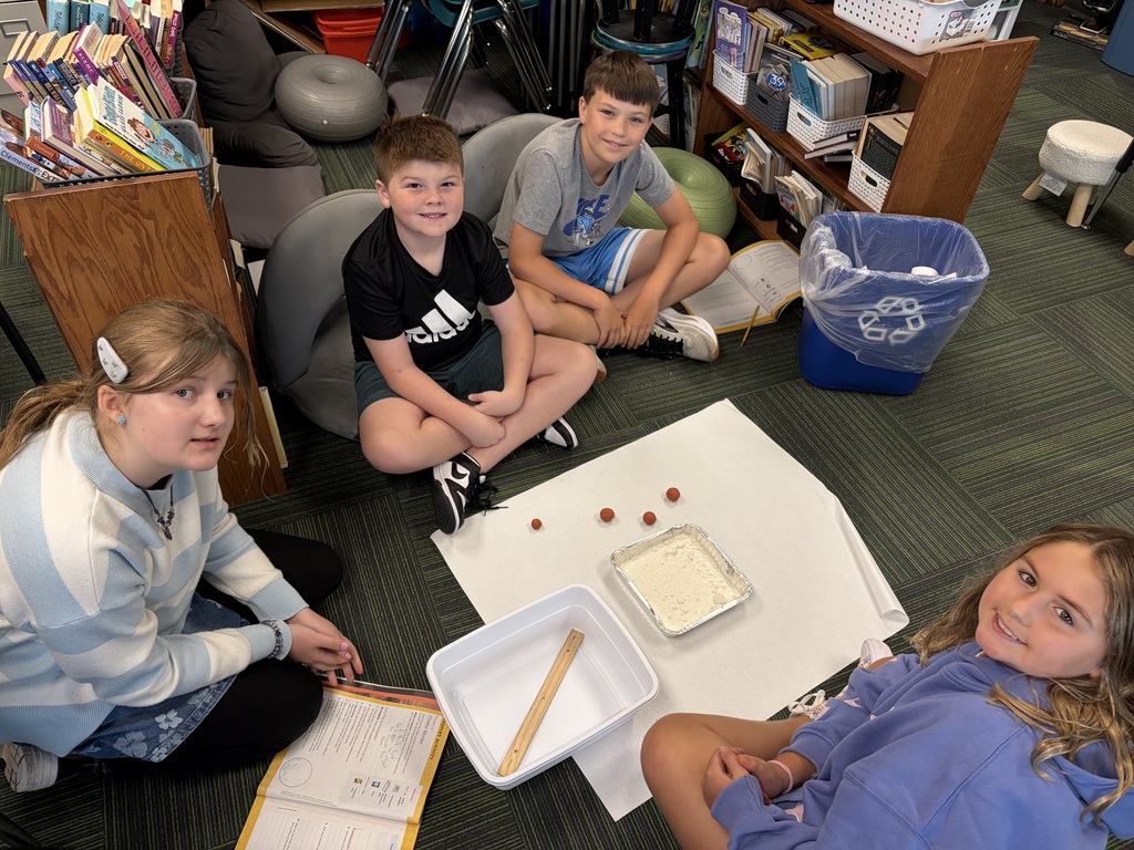 Four students seated on the carpet around a science experiment station with a tray of flour, wooden ruler, and small clay balls.