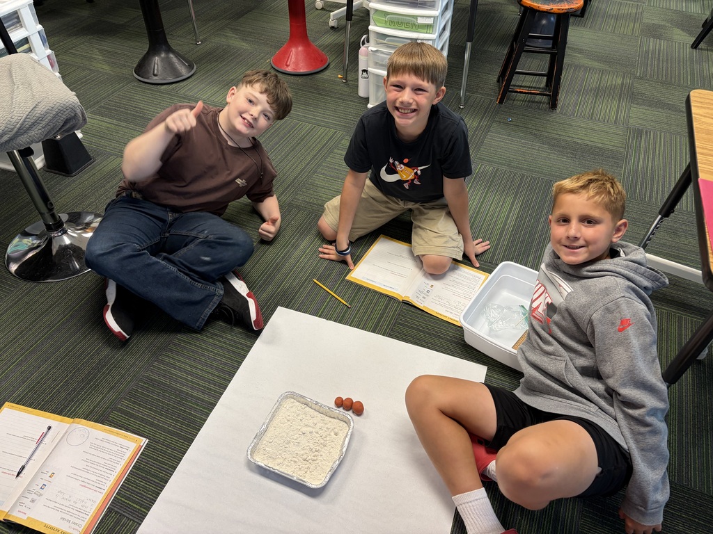 Three boys sitting around a white paper layout on the floor with open workbooks, a pan of flour, and small clay models.