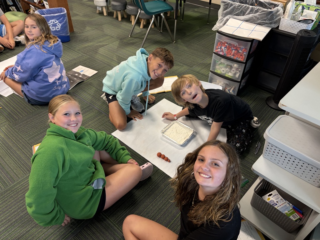 Four students sitting in a circle on the floor working on a science experiment involving a pan of flour and small clay spheres set up on paper.